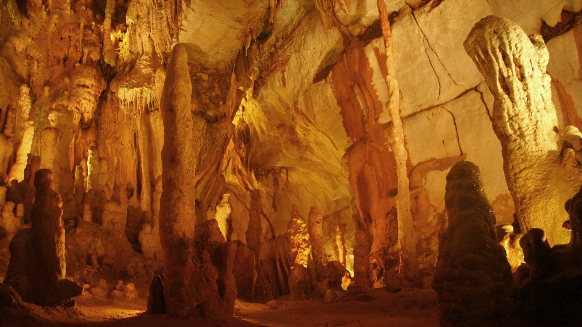 Caverna iluminada por luz dourada, com altas estalactites e estalagmites projetando sombras dramáticas.
