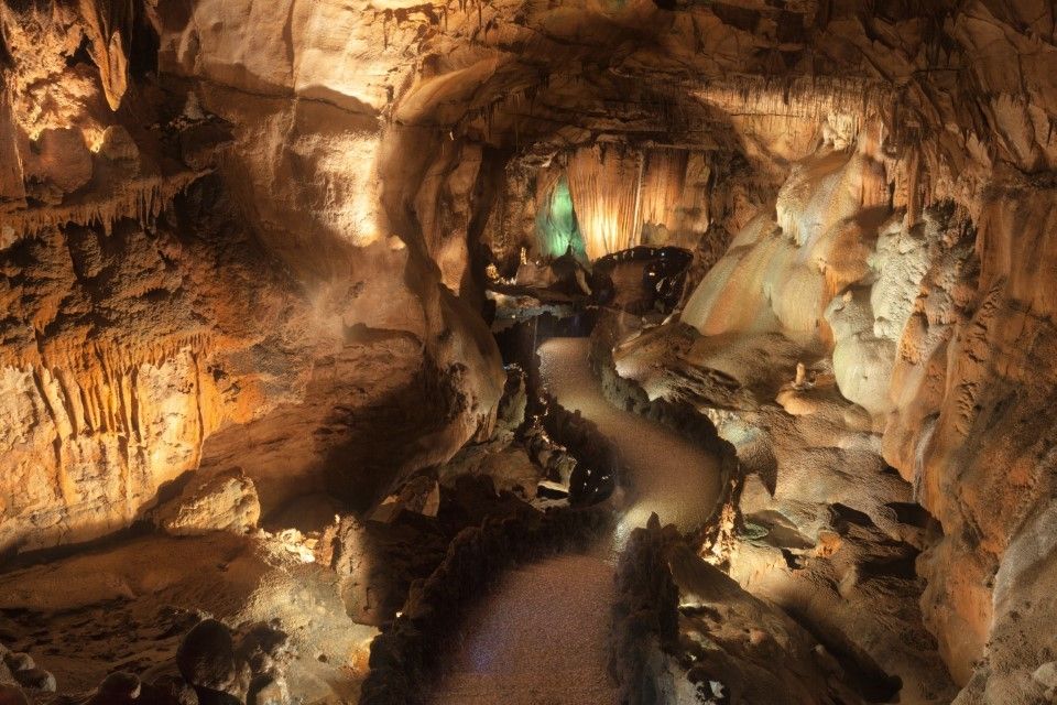Gruta estreita e iluminada com um caminho sinuoso e paredes rochosas de um laranja quente, iluminadas por uma luz verde ao longe.
