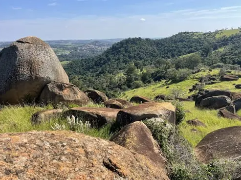 Encosta rochosa verdejante com grandes pedras, debruçada sobre um vale distante sob um céu límpido.