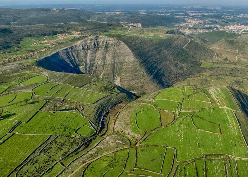 Vista aérea de uma cratera vulcânica verde com campos em terraços e uma paisagem florestal ao fundo.
