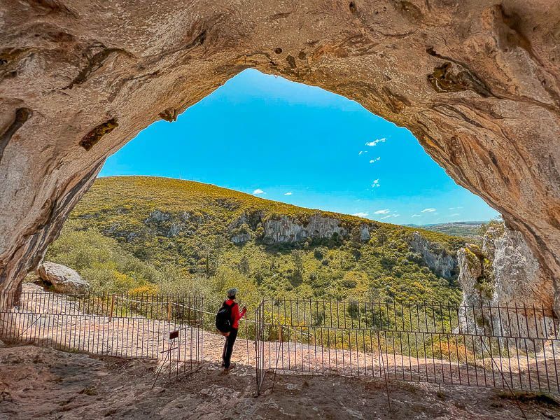 Pessoa em pé na entrada de uma caverna, olhando para uma paisagem montanhosa ensolarada.