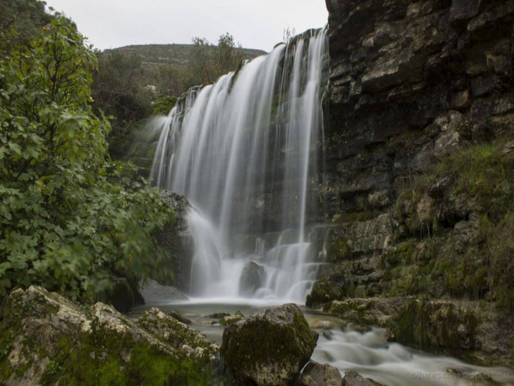 Cachoeira que deságua em uma piscina rochosa ao lado de um penhasco, emoldurada por folhagem verde.