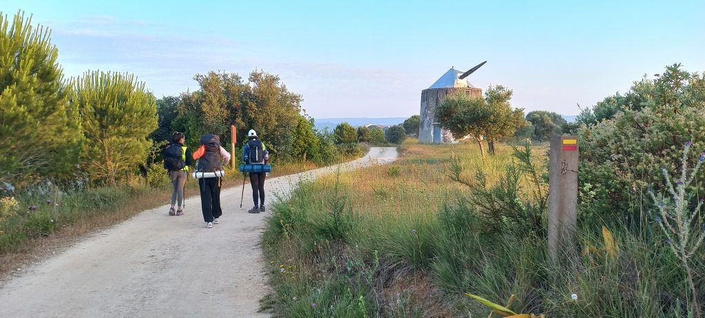 Duas pessoas caminhando por uma trilha de terra ao lado de arbustos e um campo ao pôr do sol, com um moinho de vento ao longe.