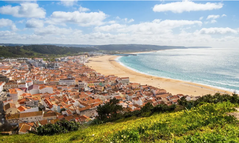 Cidade costeira com telhados de terracota ao lado de uma ampla praia de areia e oceano azul sob um céu parcialmente nublado.