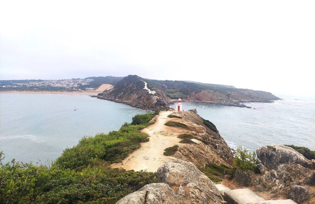 Trilha costeira rochosa que leva a uma península estreita, com o mar em ambos os lados e um pequeno farol vermelho à frente.