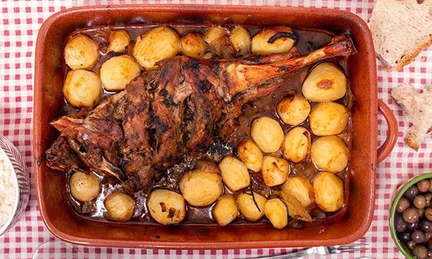 Perna de cordeiro assada com batatas em uma assadeira vermelha sobre uma toalha de mesa xadrez.
