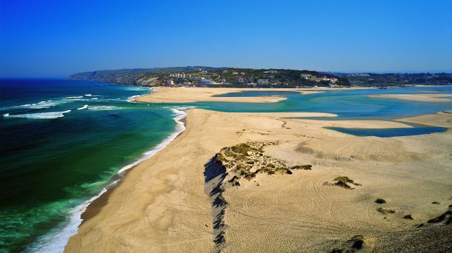 Praia de areia e oceano turquesa com ondas, costa rochosa e litoral distante sob um céu azul claro.