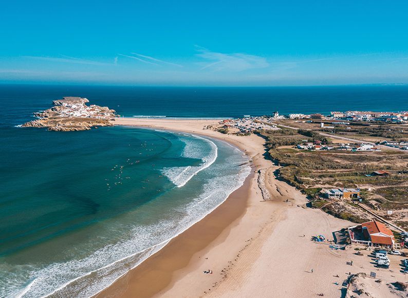 Vista aérea de uma baía turquesa, praia de areia e uma pequena vila costeira sob um céu azul.