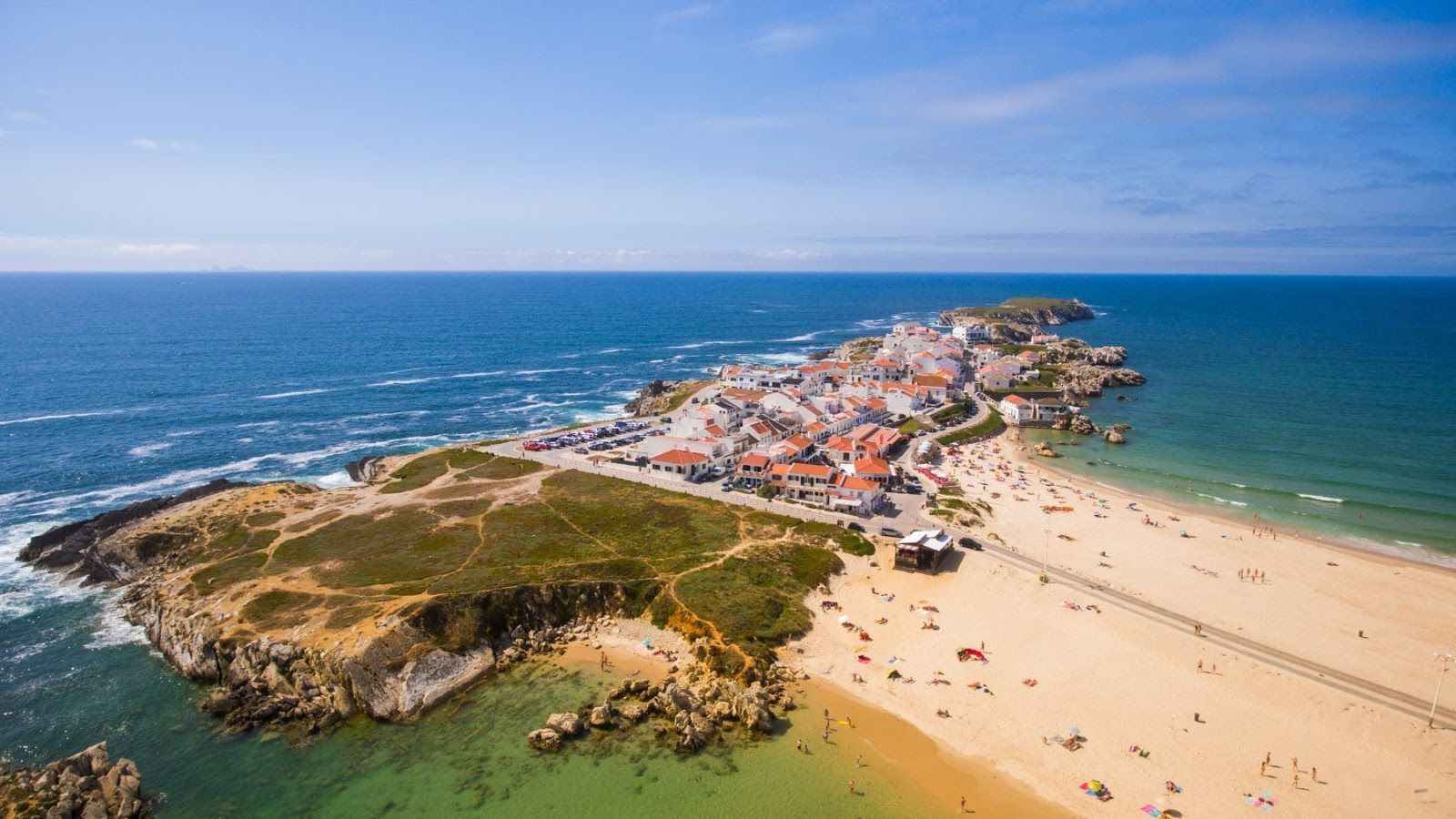 Vista aérea de uma praia de areia e cidade costeira junto ao oceano azul sob um céu limpo.