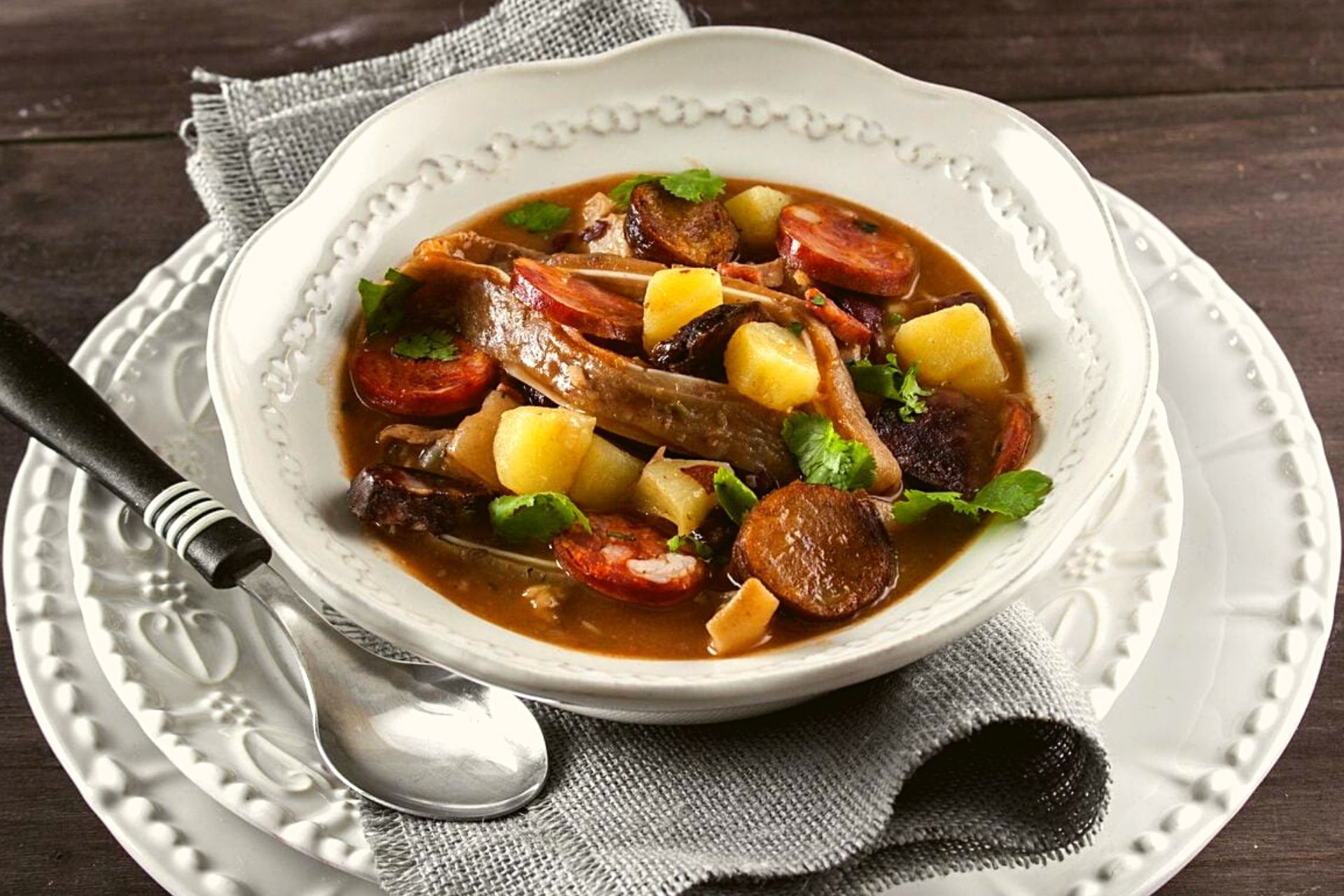Uma tigela de ensopado de carne substancioso com batatas e legumes, servida num prato branco, acompanhada de uma colher e um guardanapo.