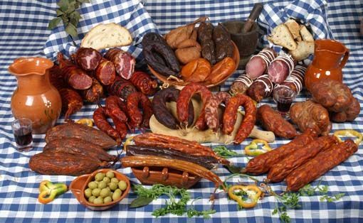 Variedade de carnes curadas e linguiças dispostas sobre uma toalha de mesa xadrez azul, acompanhadas de pão e azeitonas.