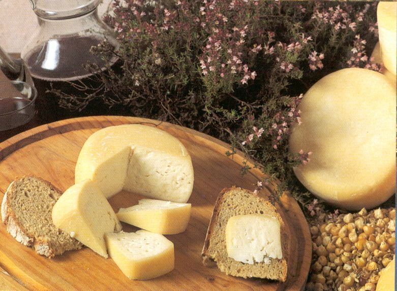 Fatias de queijo e uma roda inteira numa tábua de madeira com flores e nozes por perto.