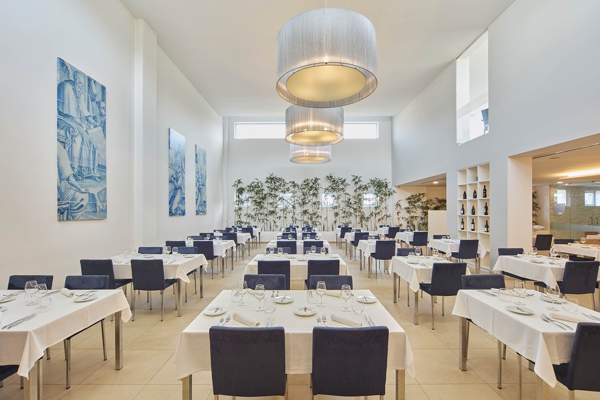 Sala de jantar moderna e iluminada com mesas brancas, cadeiras azul-marinho e grandes luminárias pendentes.