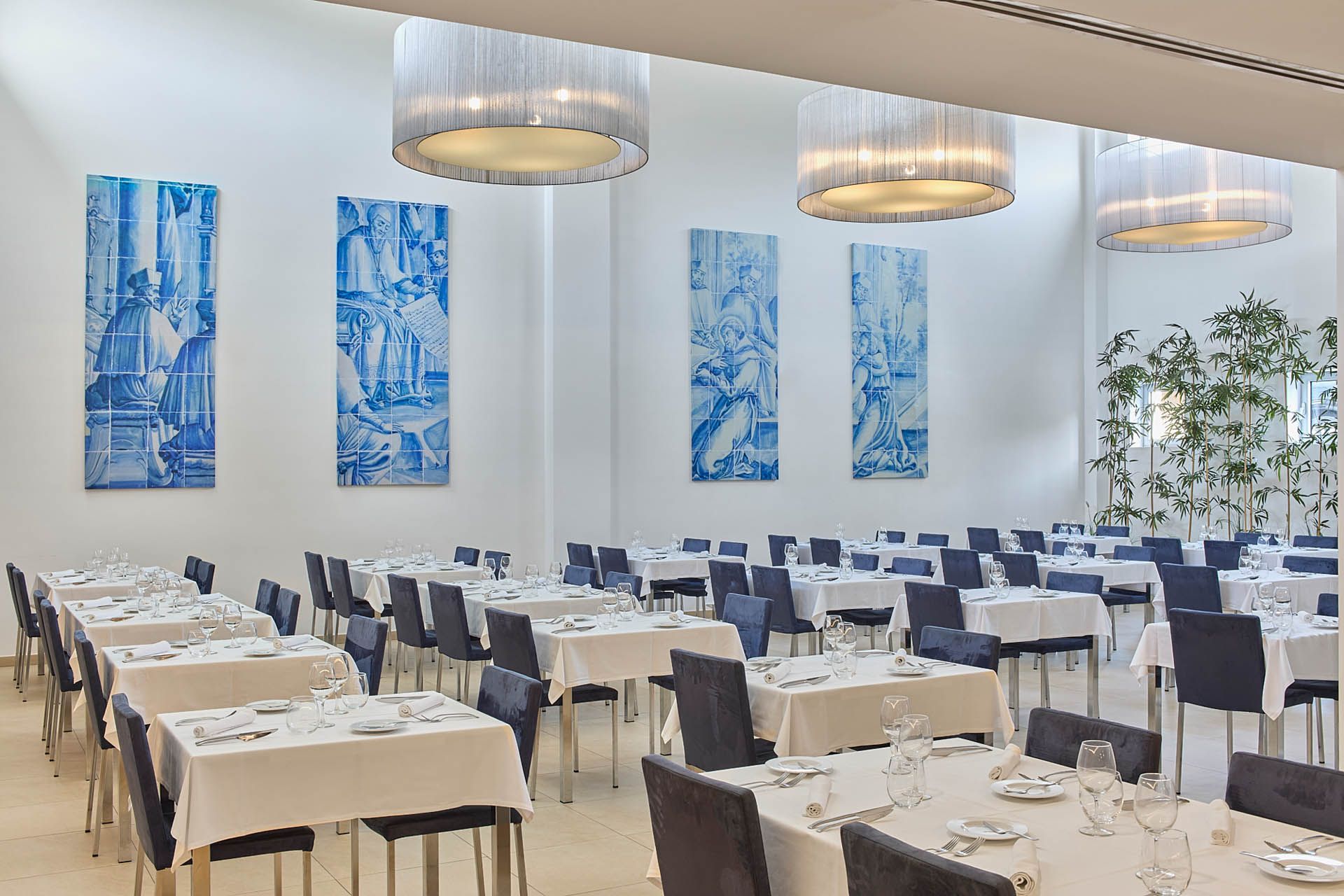 Salão de banquetes moderno e luminoso com fileiras de mesas postas, cadeiras azul-marinho, obras de arte azuis nas paredes e luminárias pendentes.