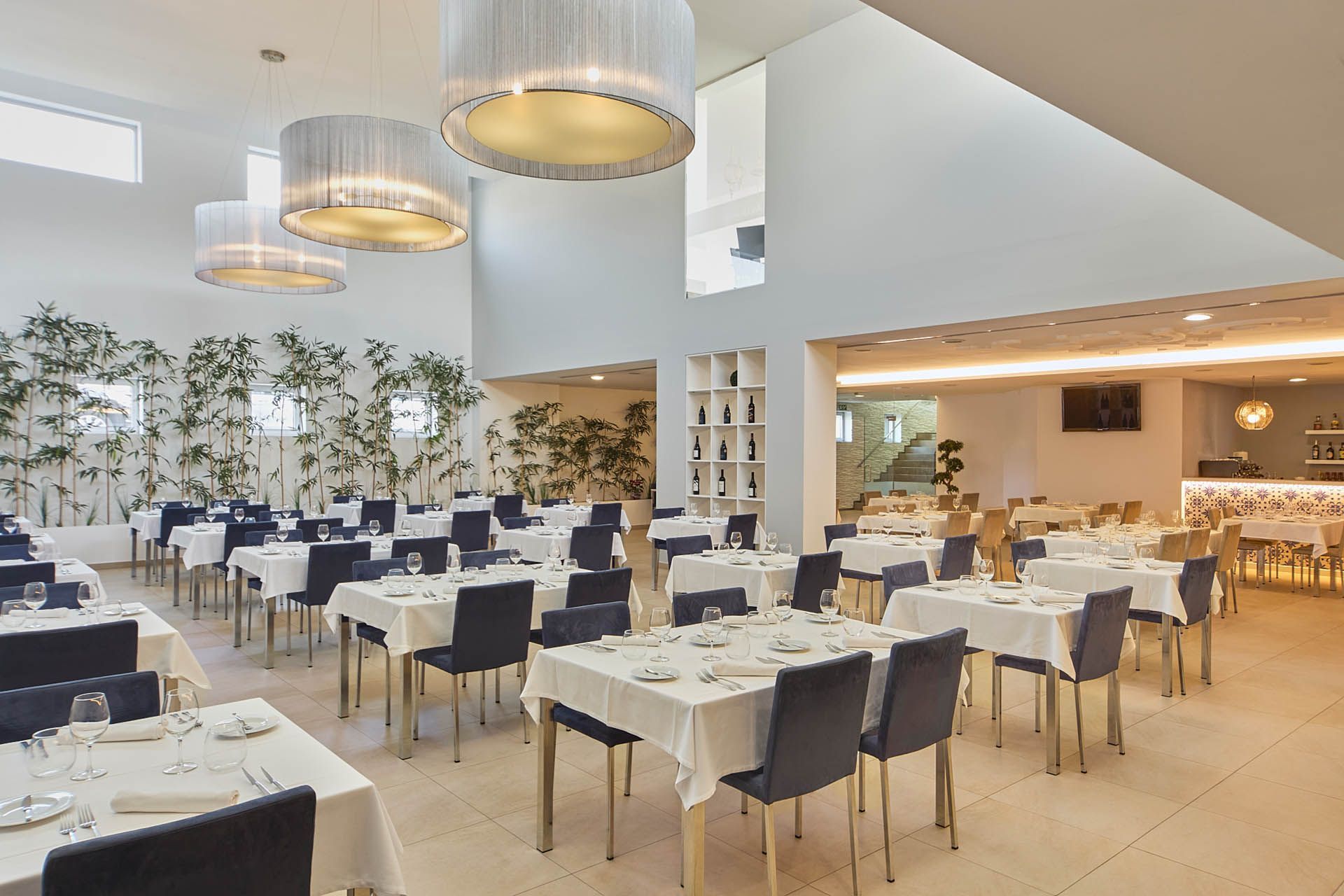 Sala de jantar de restaurante moderna e iluminada, com mesas postas, cadeiras pretas e luminárias pendentes.