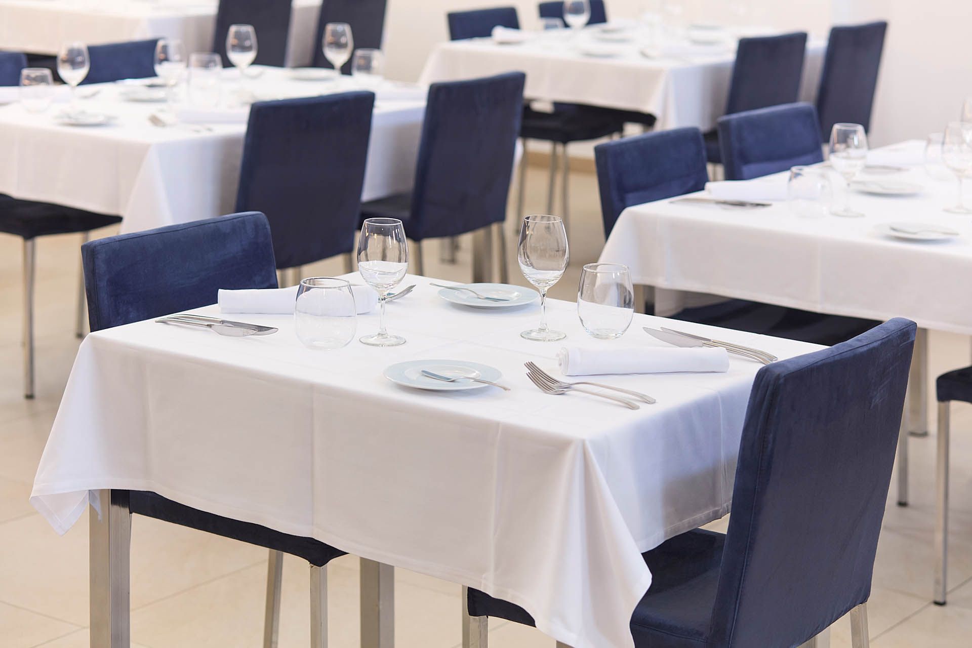 Área de jantar de restaurante vazia com toalhas de mesa brancas, cadeiras azuis e mesas postas.