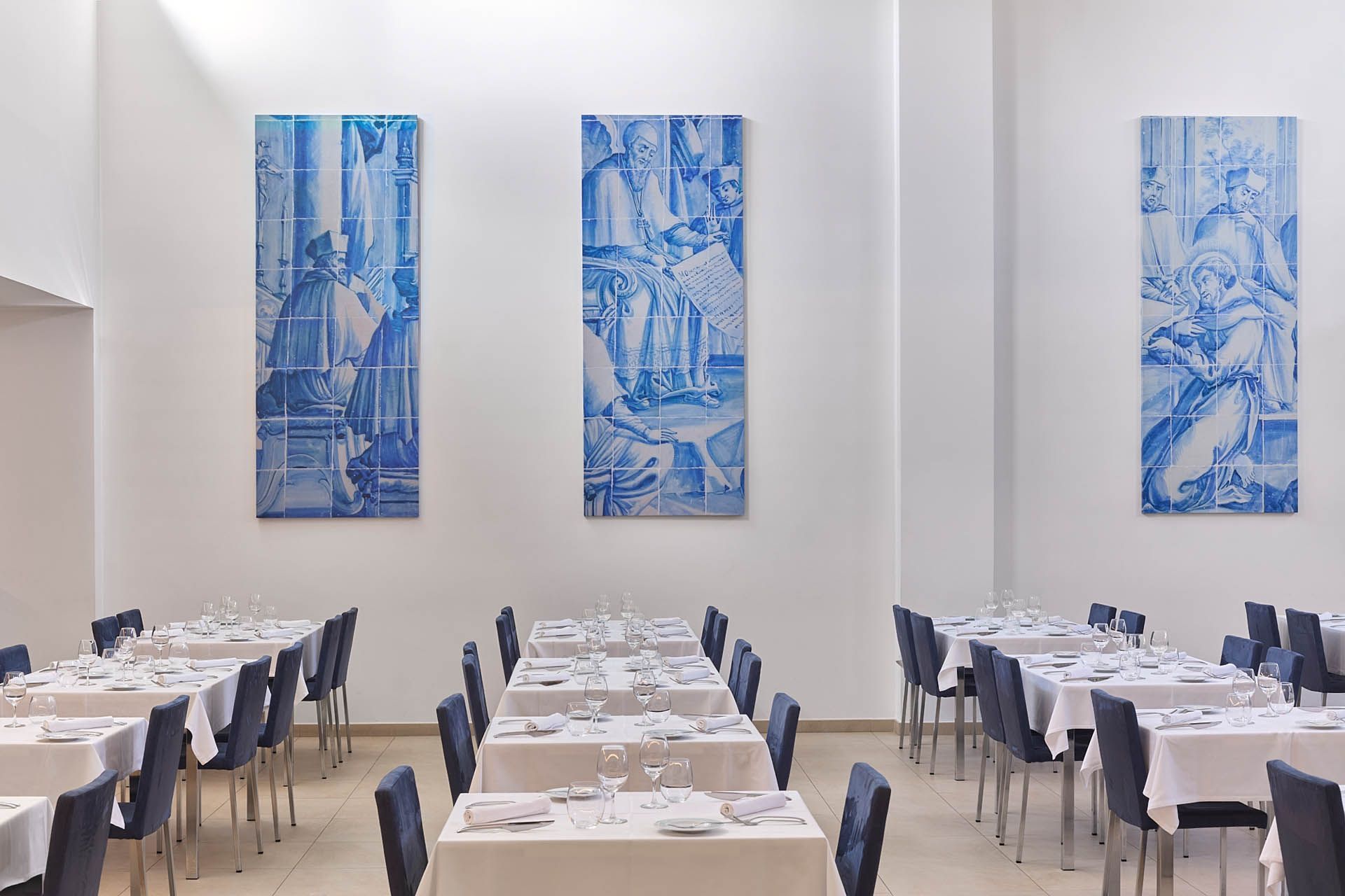 Sala de jantar moderna com toalhas de mesa brancas, cadeiras escuras e obras de arte emolduradas em azul sobre paredes creme.