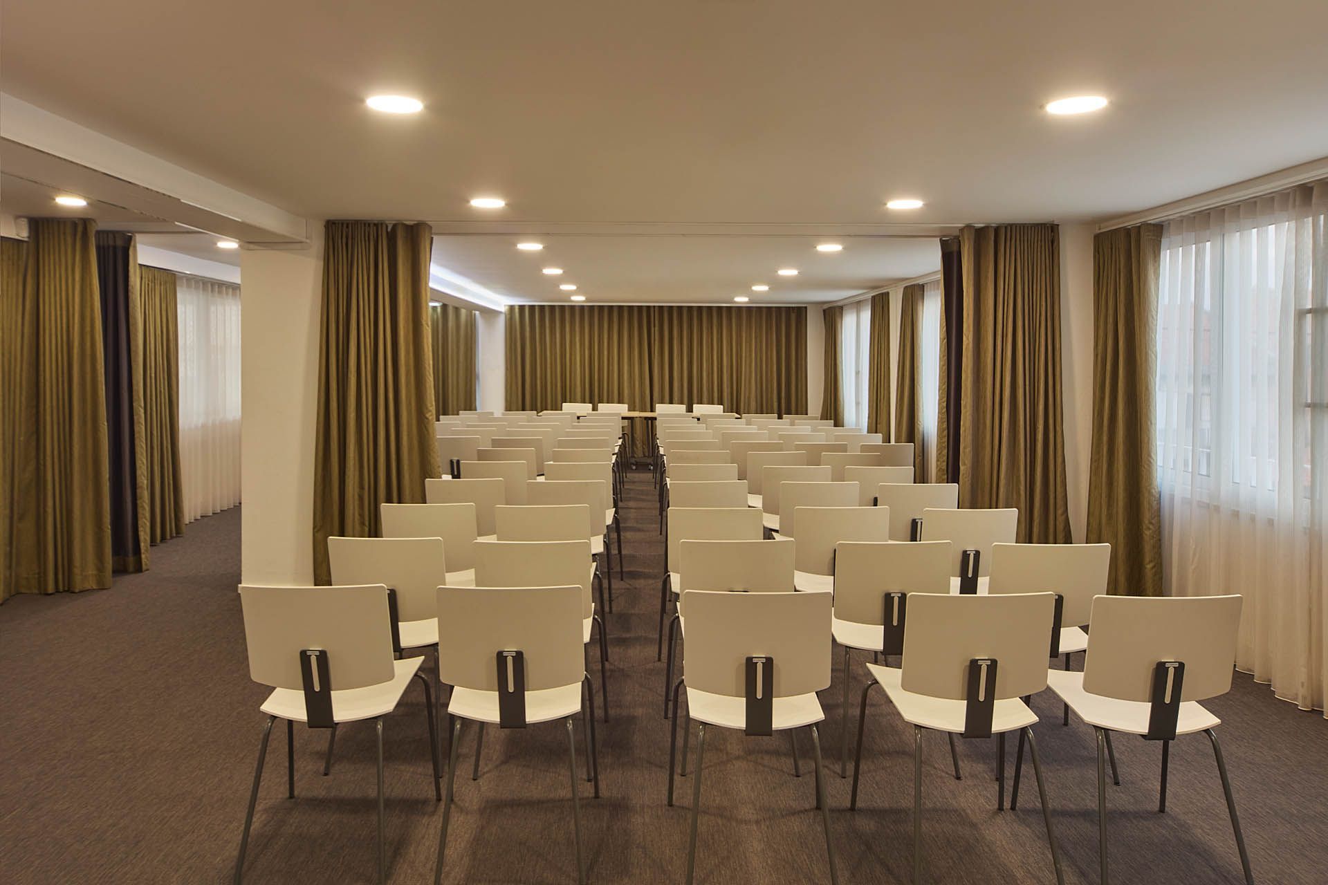 Sala de conferências vazia com fileiras de cadeiras bege, piso acarpetado e luzes no teto.