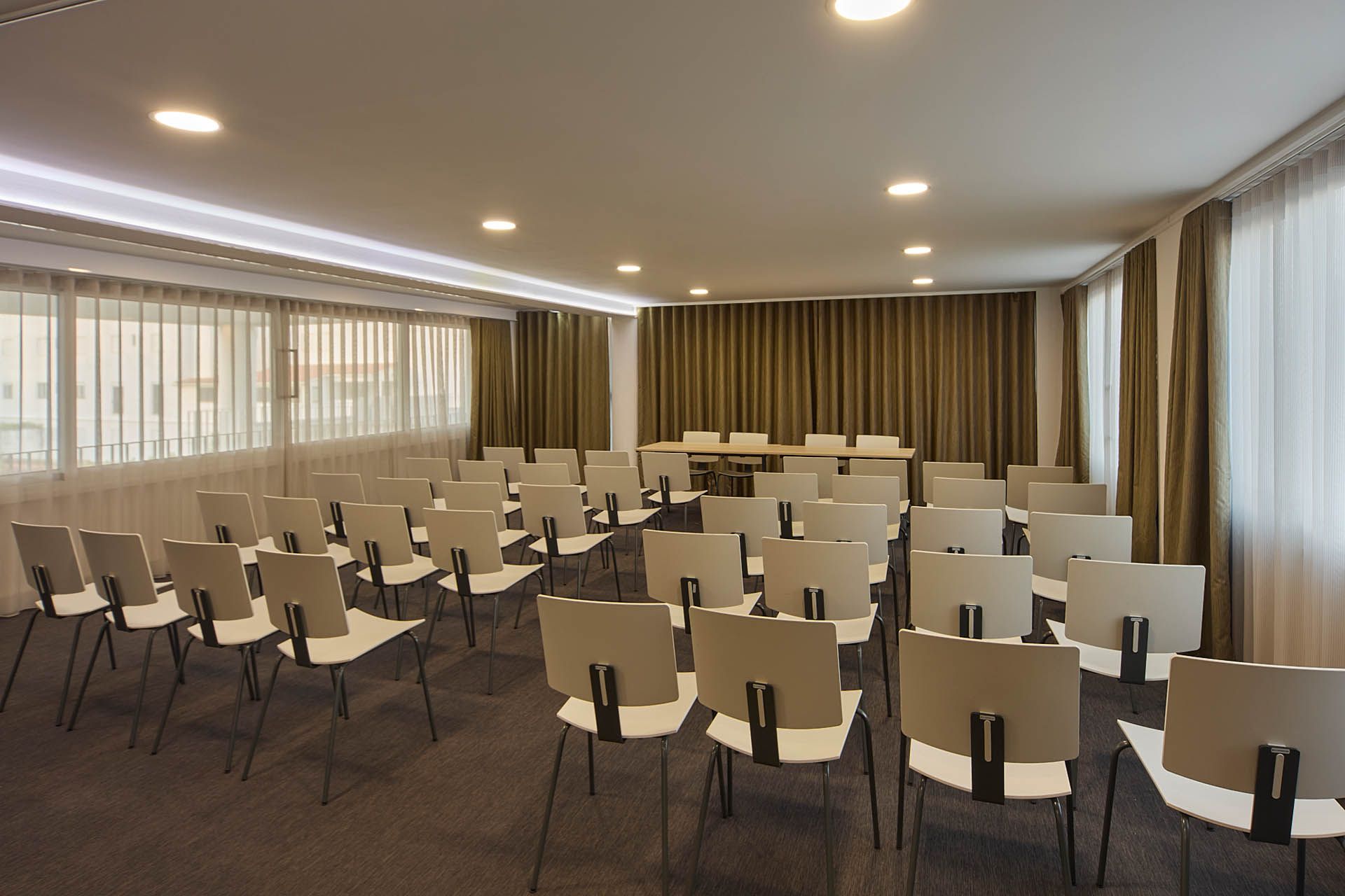 Sala de conferências vazia com fileiras de cadeiras bege voltadas para uma área de apresentação e grandes janelas com cortinas.