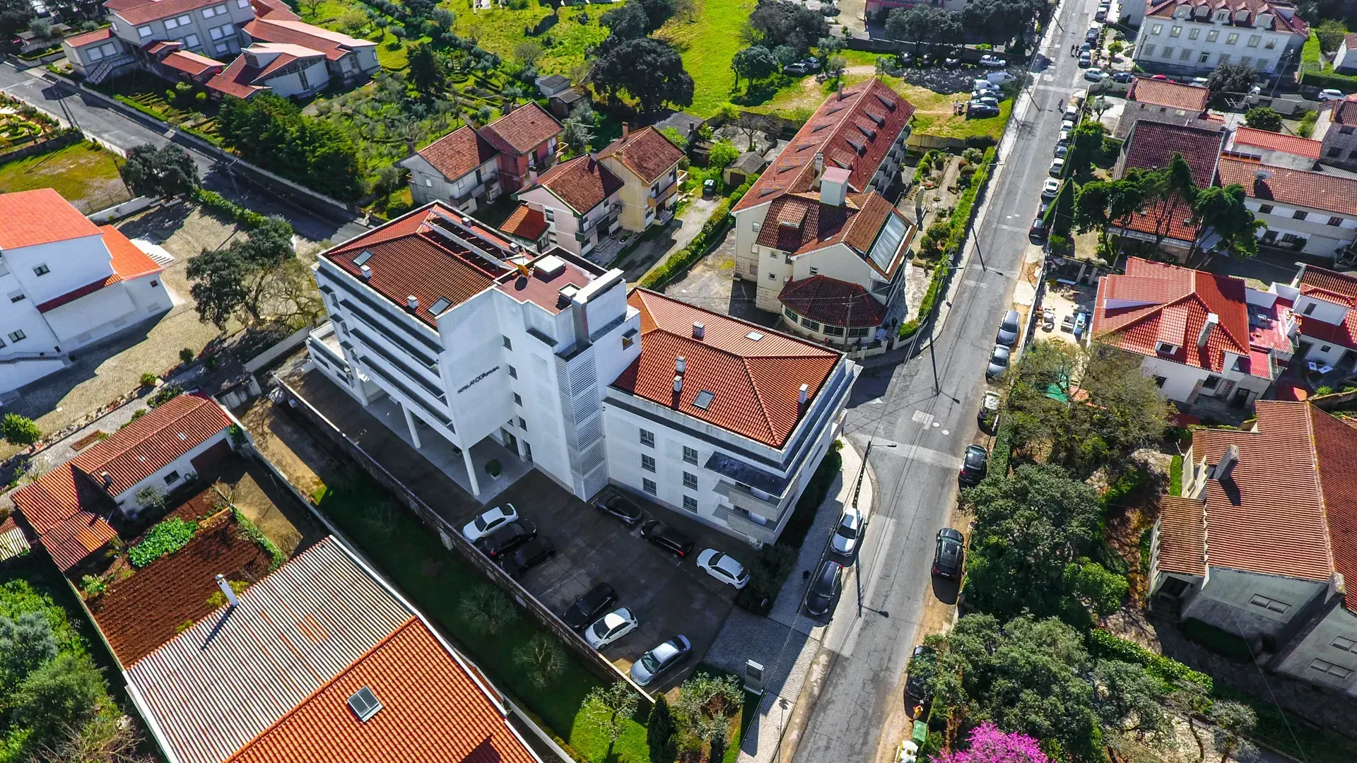 Vista aérea de um prédio de apartamentos branco em uma rua residencial com casas de telhado vermelho e carros estacionados.