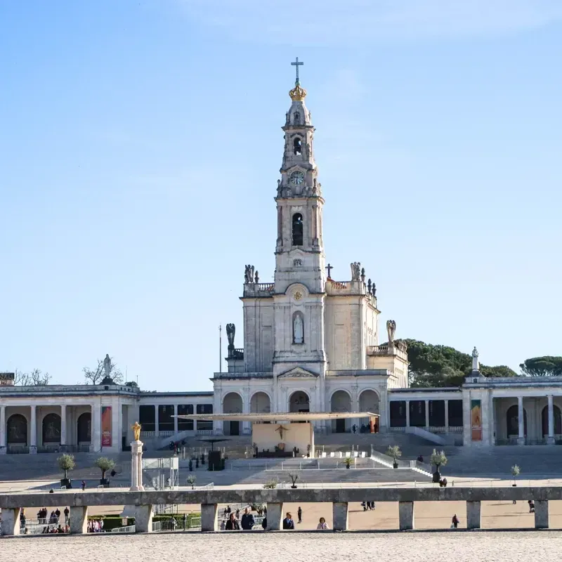 Alta basílica branca com uma torre sineira central e uma ampla praça em frente, sob um céu azul.