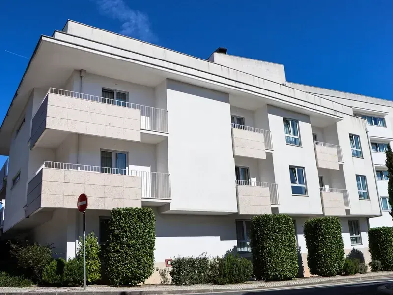 Prédio de apartamentos branco com varandas e cercas vivas sob um céu azul claro.