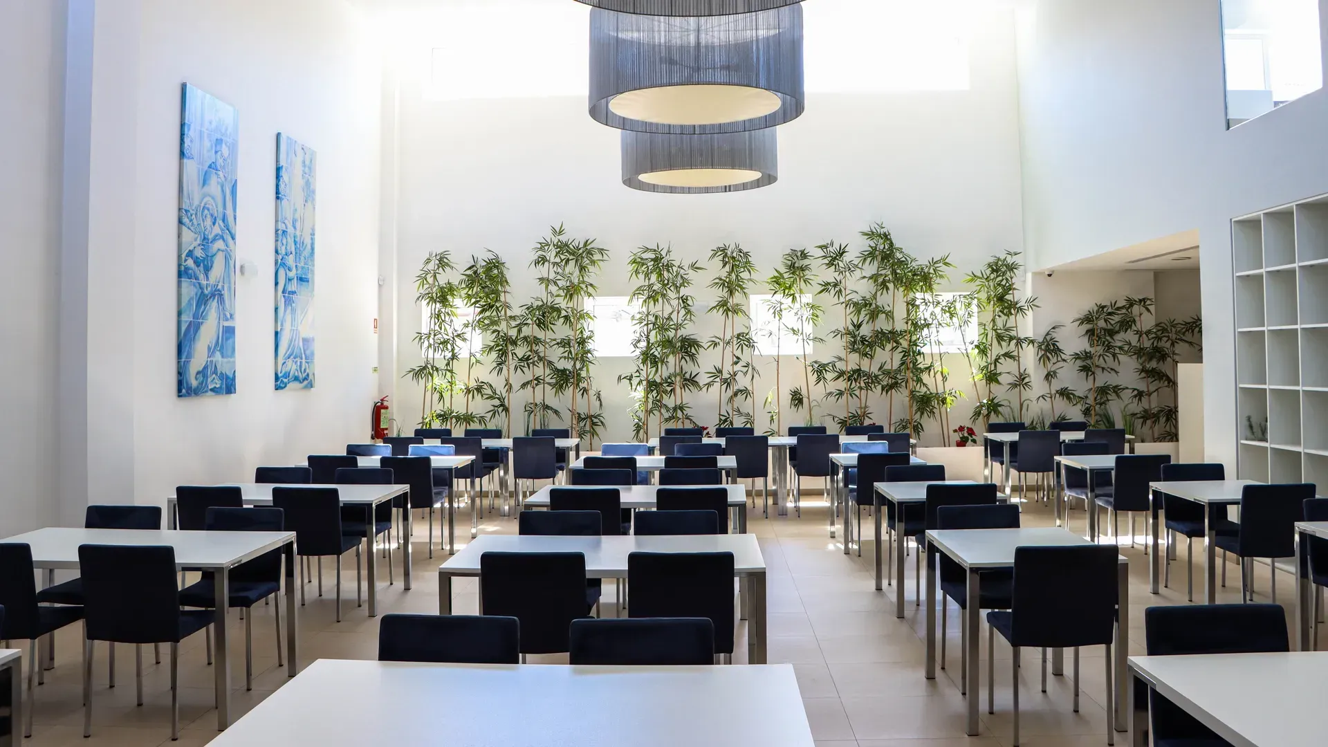 Refeitório moderno e luminoso com fileiras de mesas brancas, cadeiras pretas e grandes janelas com plantas.