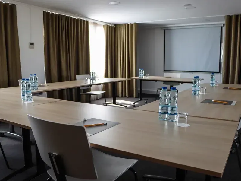 Sala de conferências vazia com uma mesa em formato de U, garrafas de água, cadeiras e uma tela de projeção.