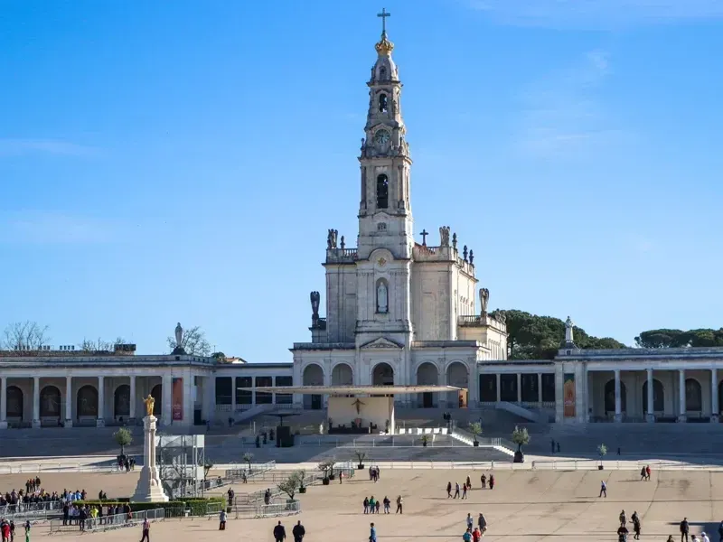 Igreja branca com campanário, debruçada sobre uma praça movimentada sob um céu azul límpido.