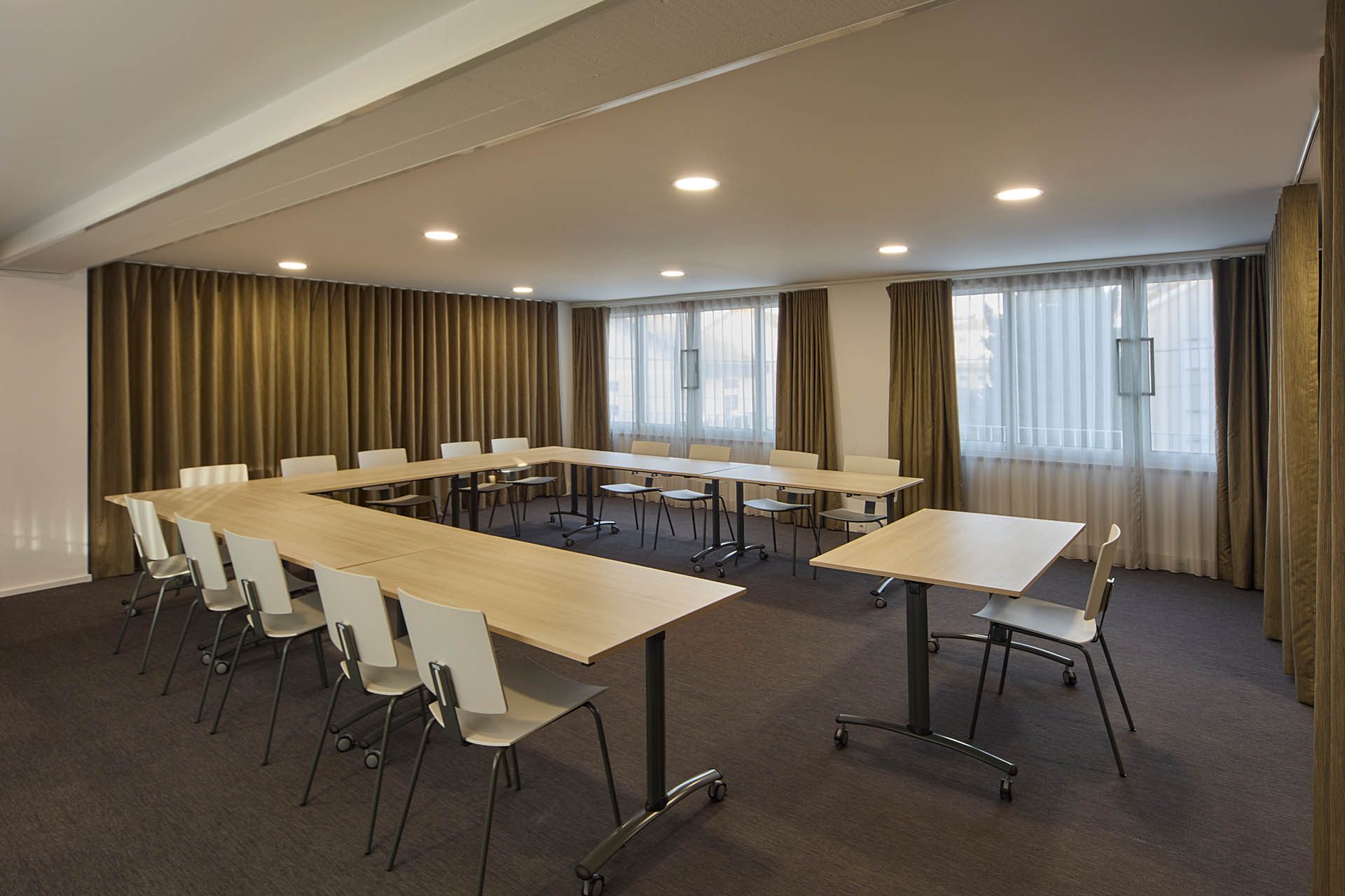 Sala de conferências vazia com mesas em formato de U, cadeiras pretas, piso acarpetado e janelas com cortinas.