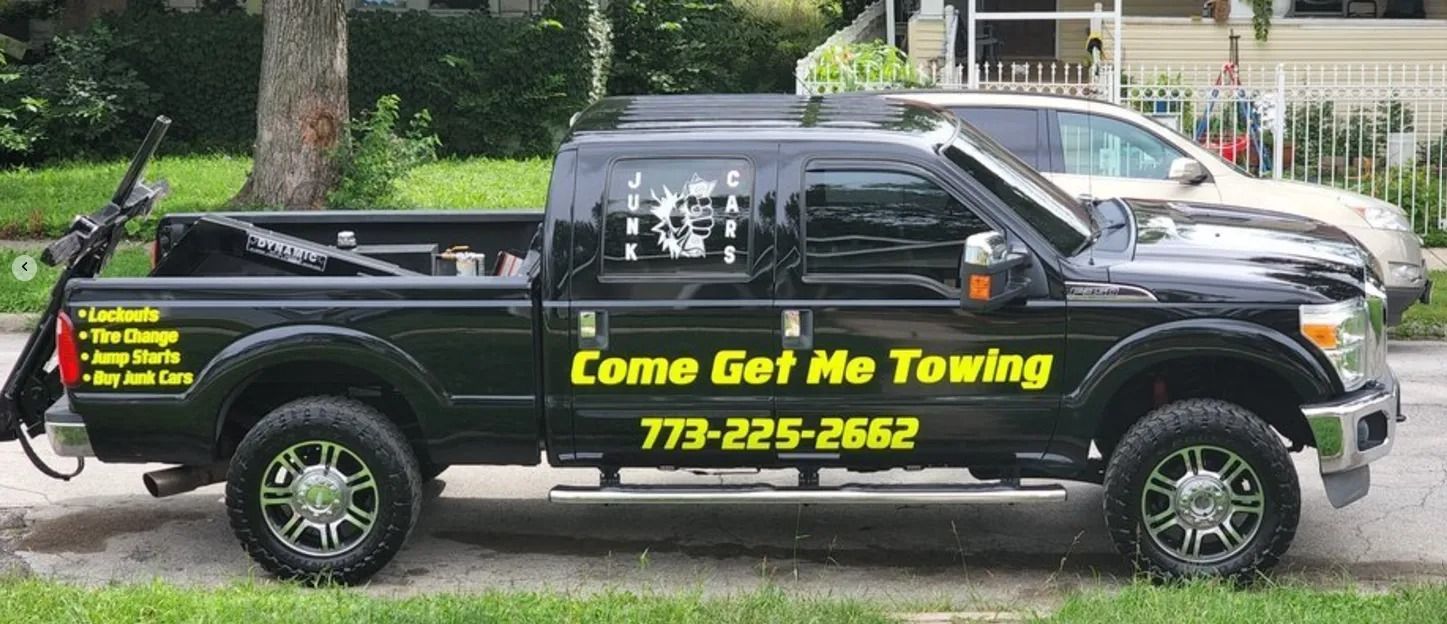 Black towing pickup truck with yellow “Come Get Me Towing” lettering parked on a street