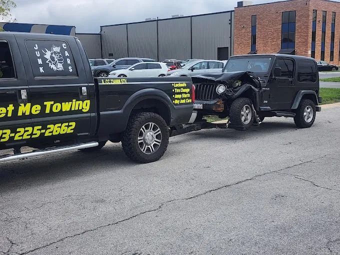 Black Jeep being towed by a black tow truck in a parking lot near a brick building.