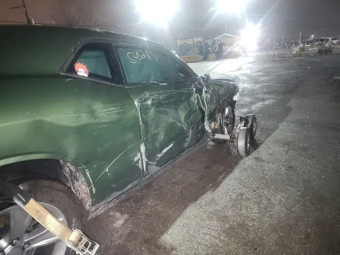 Green car with front-end damage in a dimly lit parking lot at night