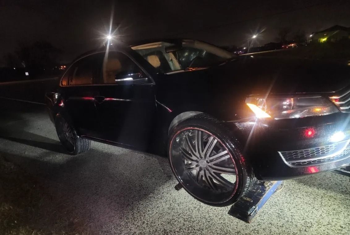 Black sedan with front wheel turned, parked on a dark street at night with headlights on.