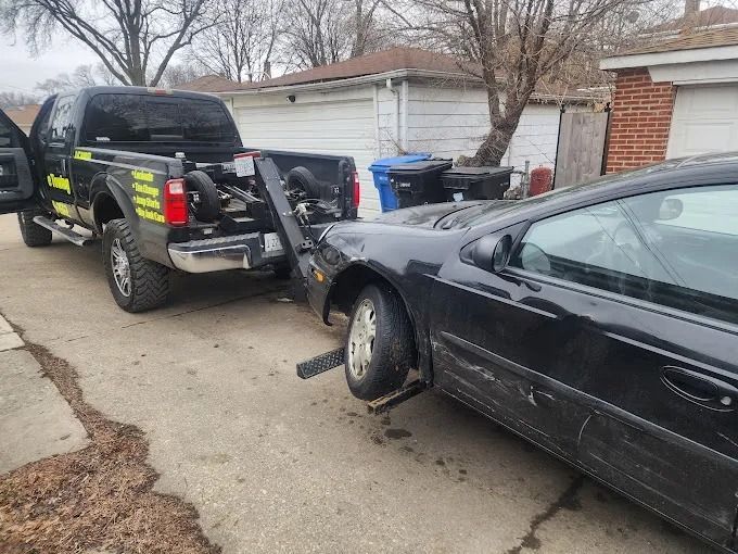 Tow truck hooked to a black car with a missing front wheel in a driveway beside a garage