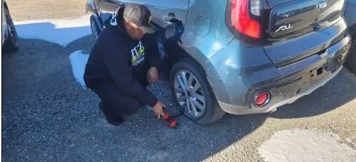 Person kneels beside a gray SUV, using a red tool near the rear wheel on a sunlit driveway.