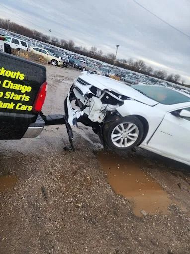 White car with severe front-end damage in a muddy parking lot near a sign and rows of vehicles