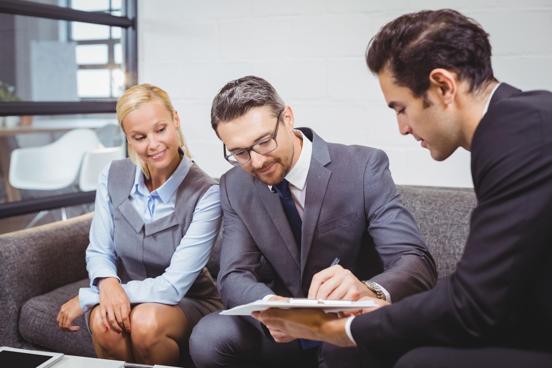 Three coworkers in business attire reviewing documents and discussing on a couch in an office lounge.