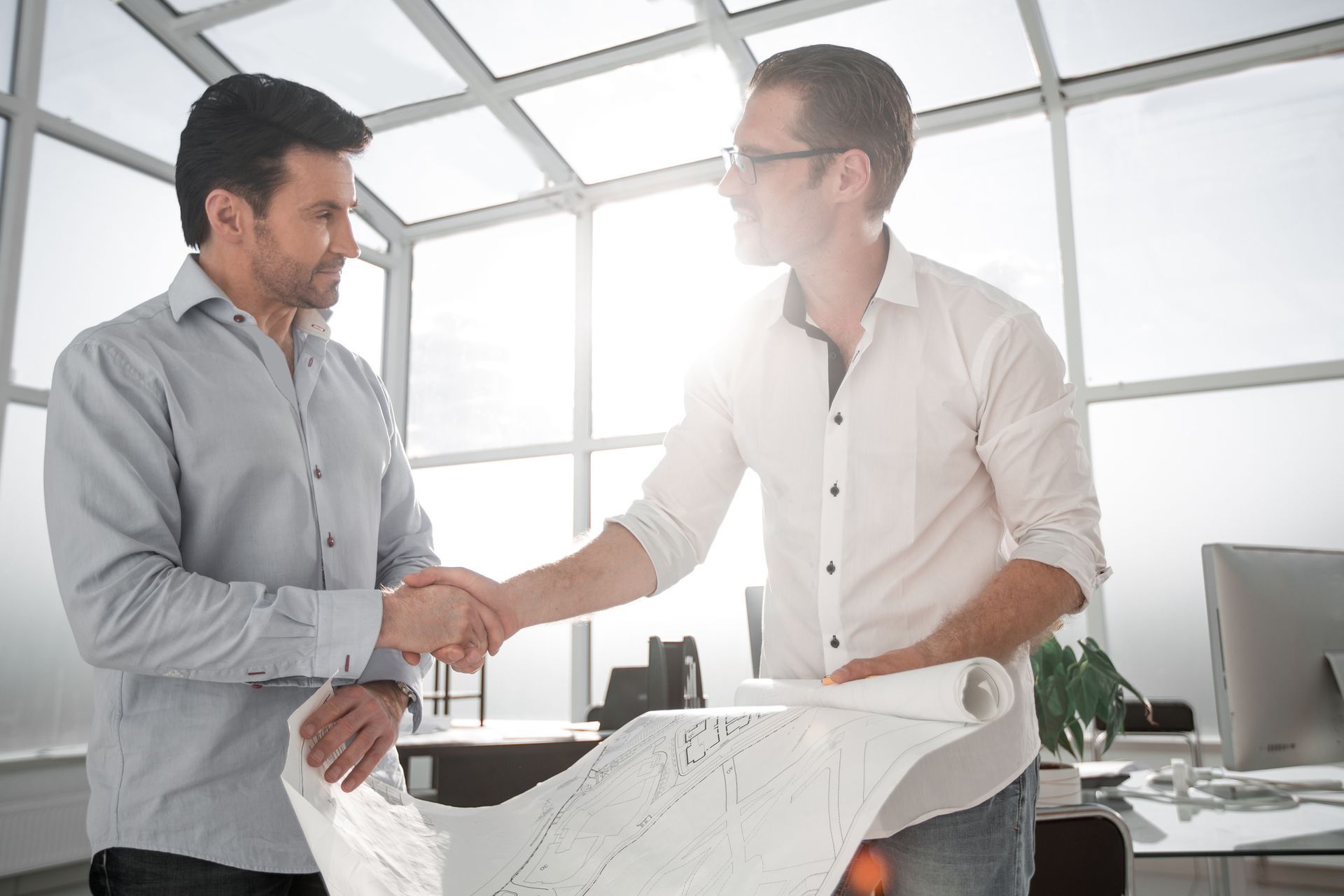 Two men shake hands in a bright office with architectural plans spread on a table.