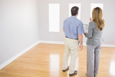 Two people standing in a bright empty room, looking at the wall and windows.