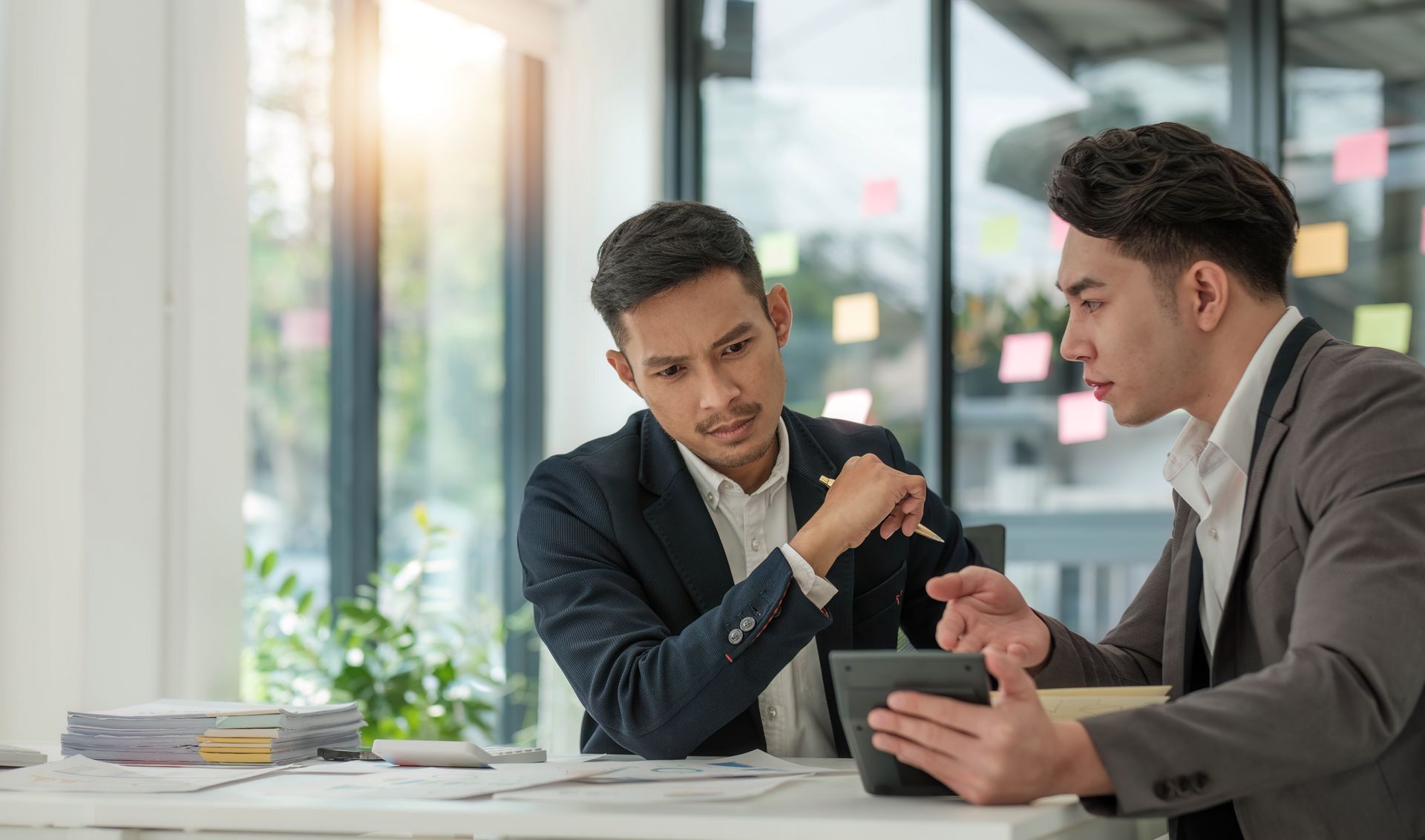 Two coworkers discuss a tablet at a desk in a bright office, with charts and papers nearby.