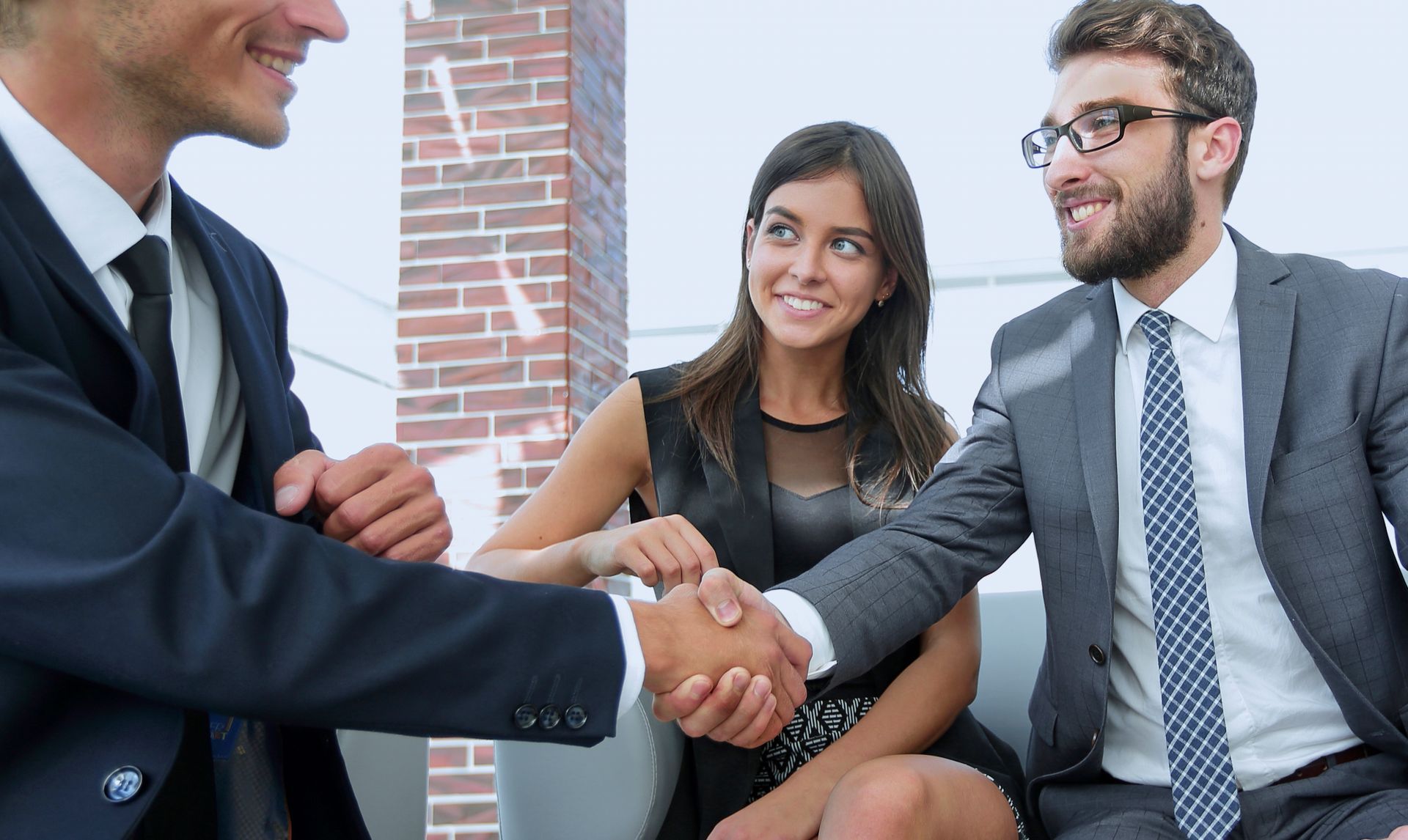 Two businesspeople shake hands in a bright office meeting with a smiling colleague nearby.