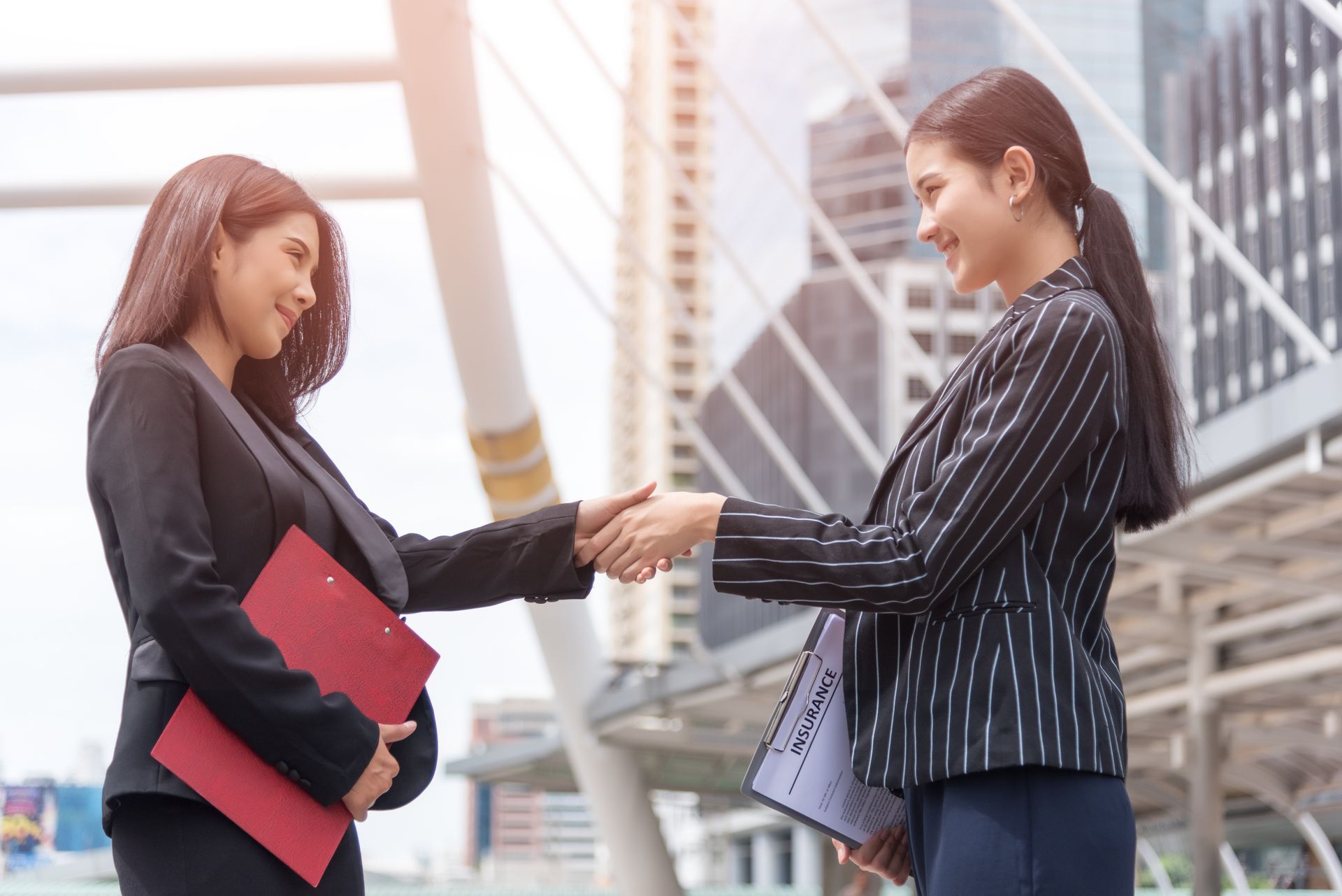 Two women shaking hands outdoors, one holding a red folder, near modern buildings.