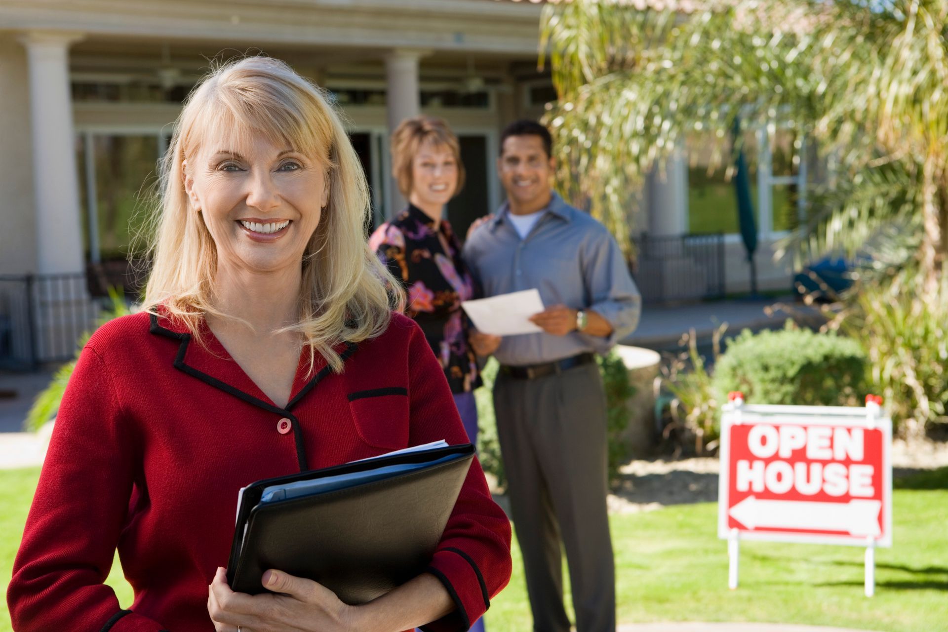 Real estate agent holding folder in front of house with two people and an open house sign