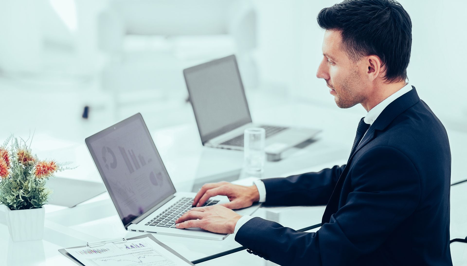 Professional man typing on a laptop at a bright office desk with two open laptops.