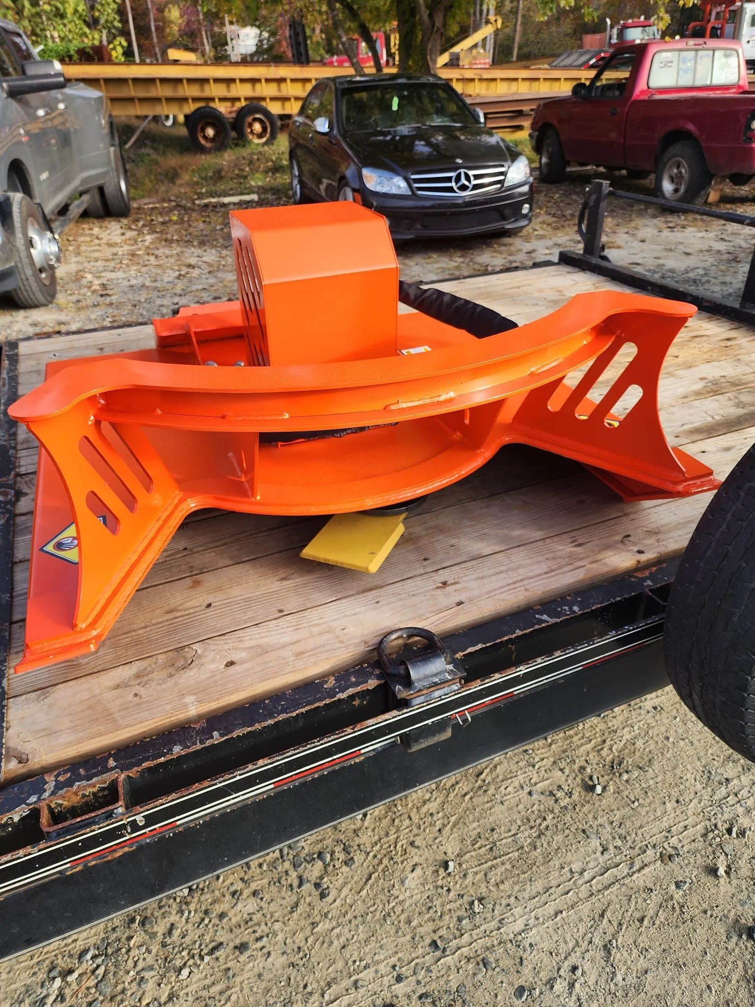 Bright orange metal machine or bench frame on a trailer in a gravel yard, with cars in the background
