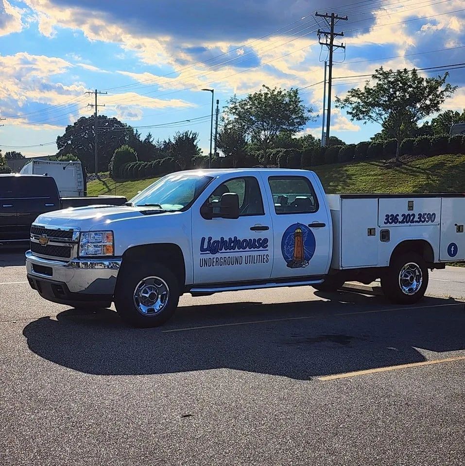 White LightHouse utility truck parked on a road under a cloudy sky, with utility trailer and pole in the background.