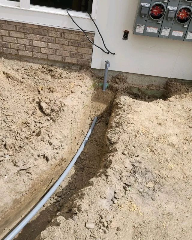 Trench beside a house with a buried gray conduit running toward electrical meters.