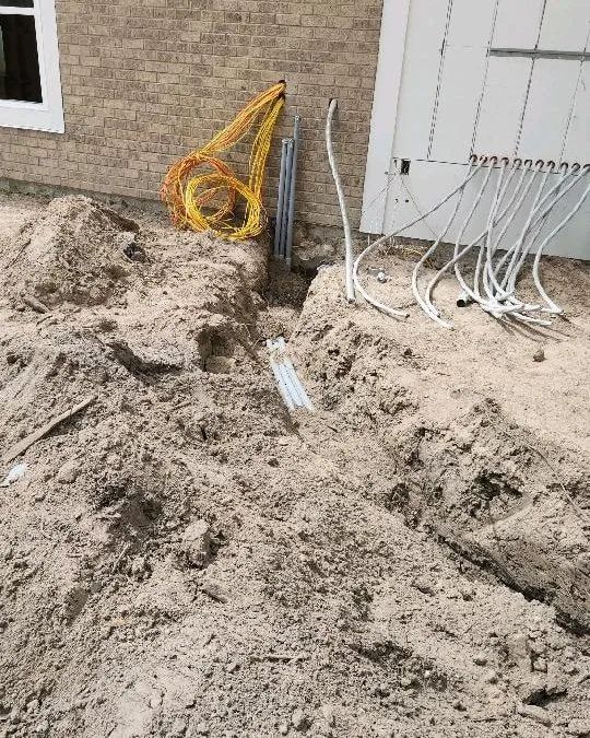 Trenches dug beside a house with exposed utility lines and sandy soil piles.