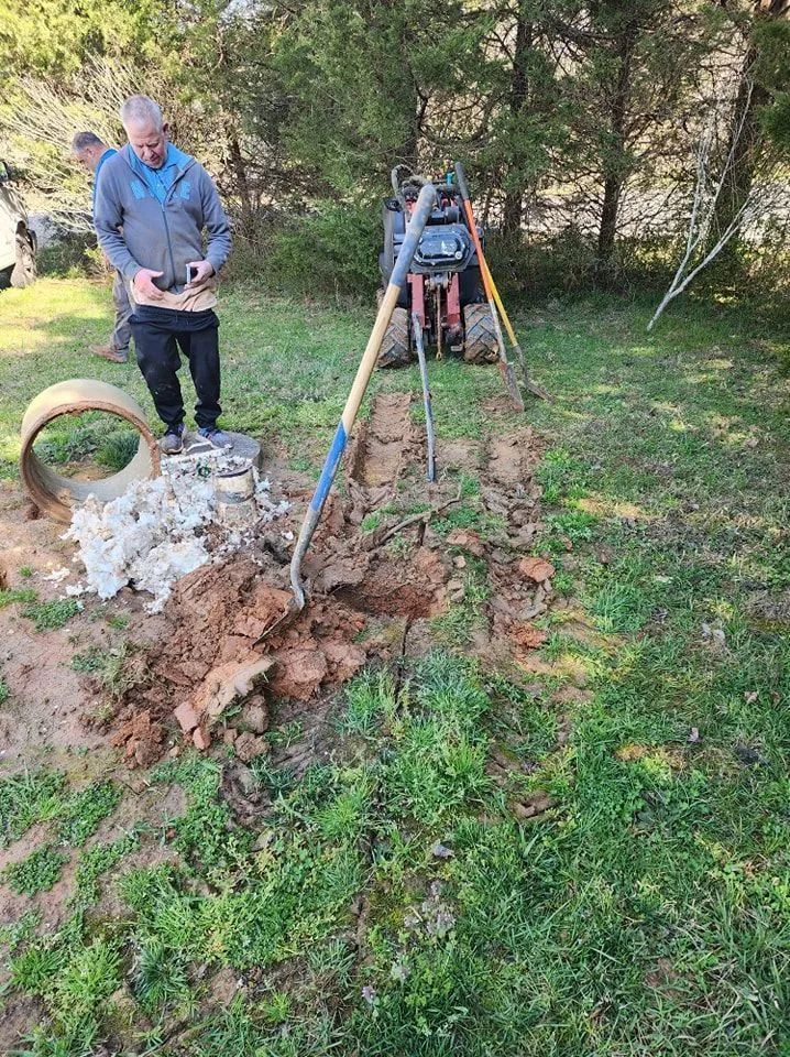 Two workers at a muddy trench beside a wooded yard, with tools and equipment nearby.